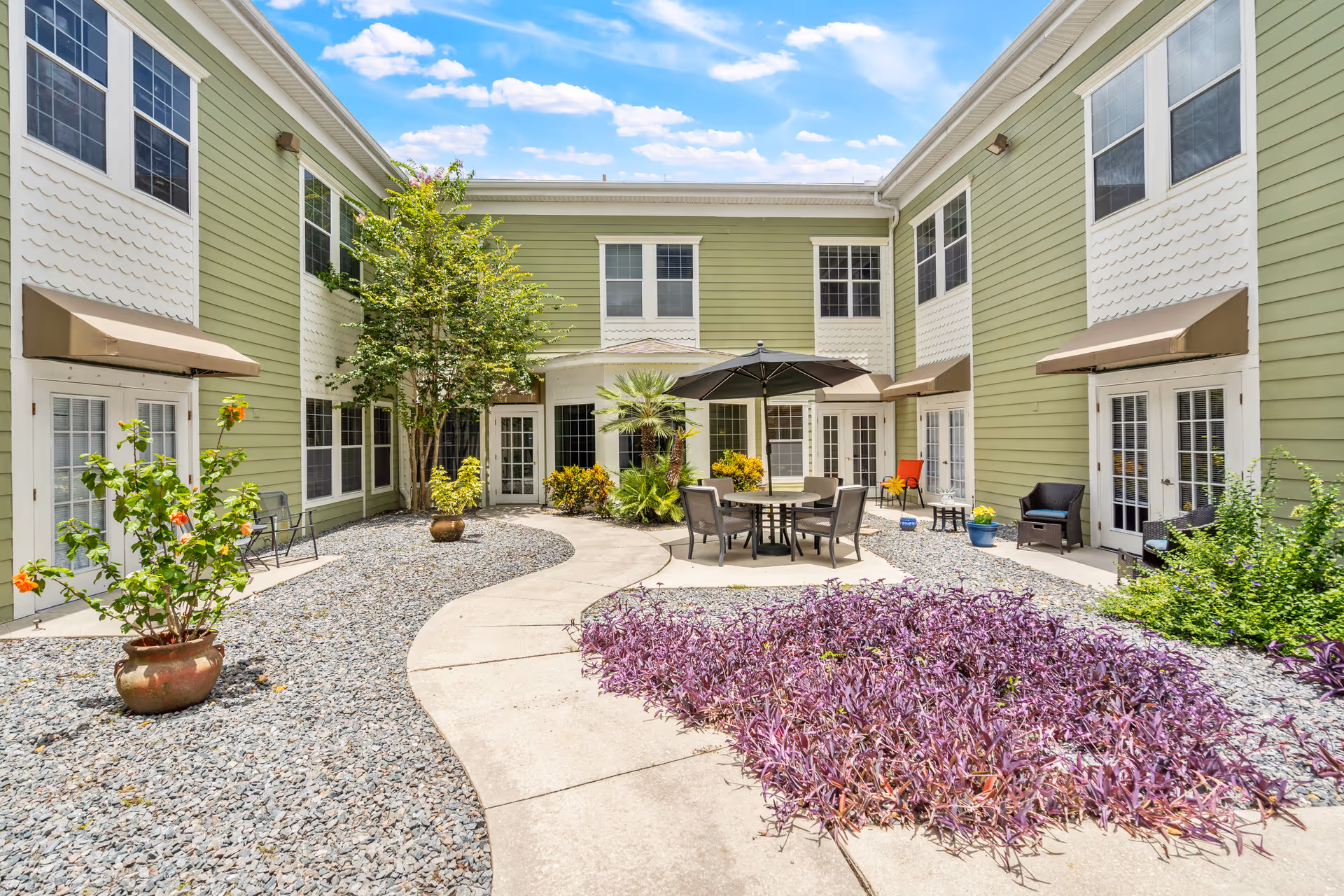 Outdoor courtyard area of a senior living facility with green and white two-story buildings surrounding a paved walkway. The courtyard features potted plants, purple ground cover, a round table with chairs and an umbrella, and additional seating along the walls under awnings. The sky is blue with some clouds.