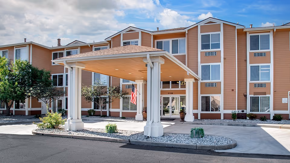 Front entrance of a three-story senior living building with a covered porte-cochere, American flag, and landscaped drive.