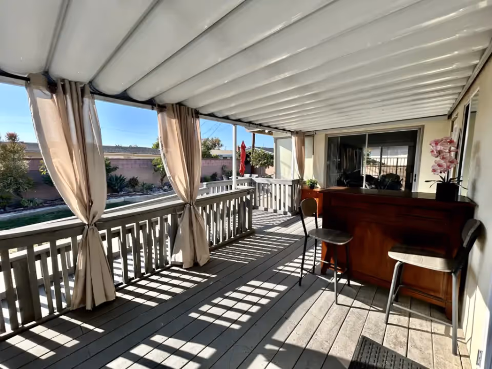 Covered outdoor patio area with a wooden floor and railing, beige curtains tied back, a wooden bar with two stools, and a potted orchid on the bar. The patio overlooks a garden with plants and a brick wall under a clear blue sky.