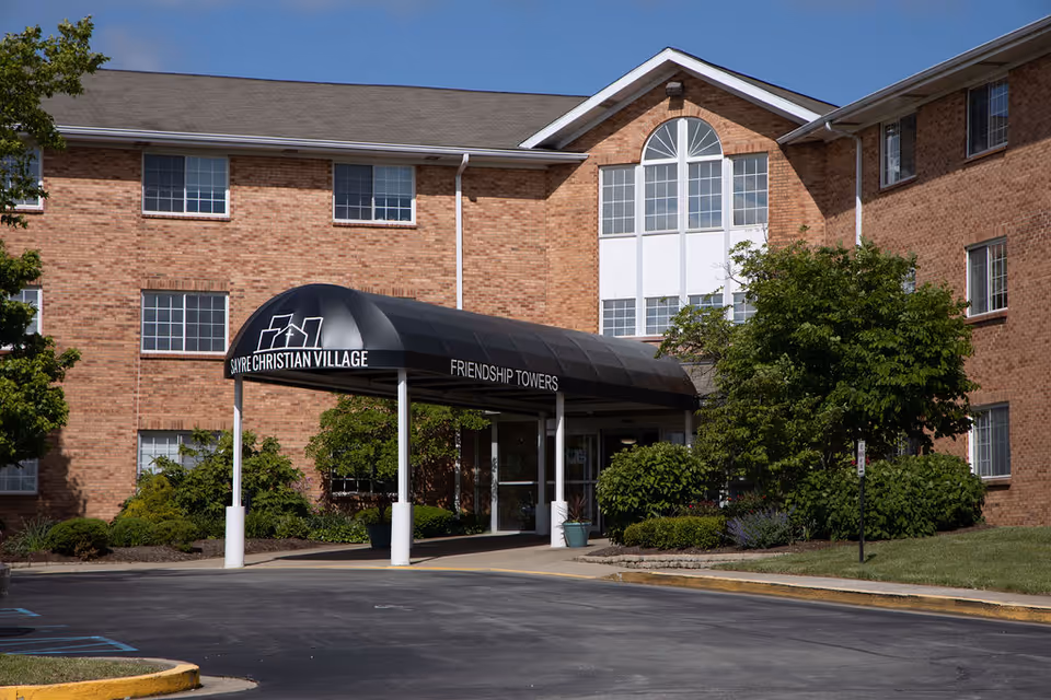 Exterior view of Sayre Christian Village building with a black awning labeled 'Sayre Christian Village Friendship Towers' over the entrance, surrounded by greenery and a clear blue sky.