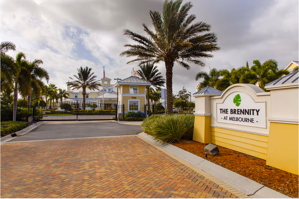 Entrance to The Brennity at Melbourne Senior Living facility with a gated driveway, palm trees, and a sign displaying the facility's name. The building in the background is yellow with white trim and has a metal roof.
