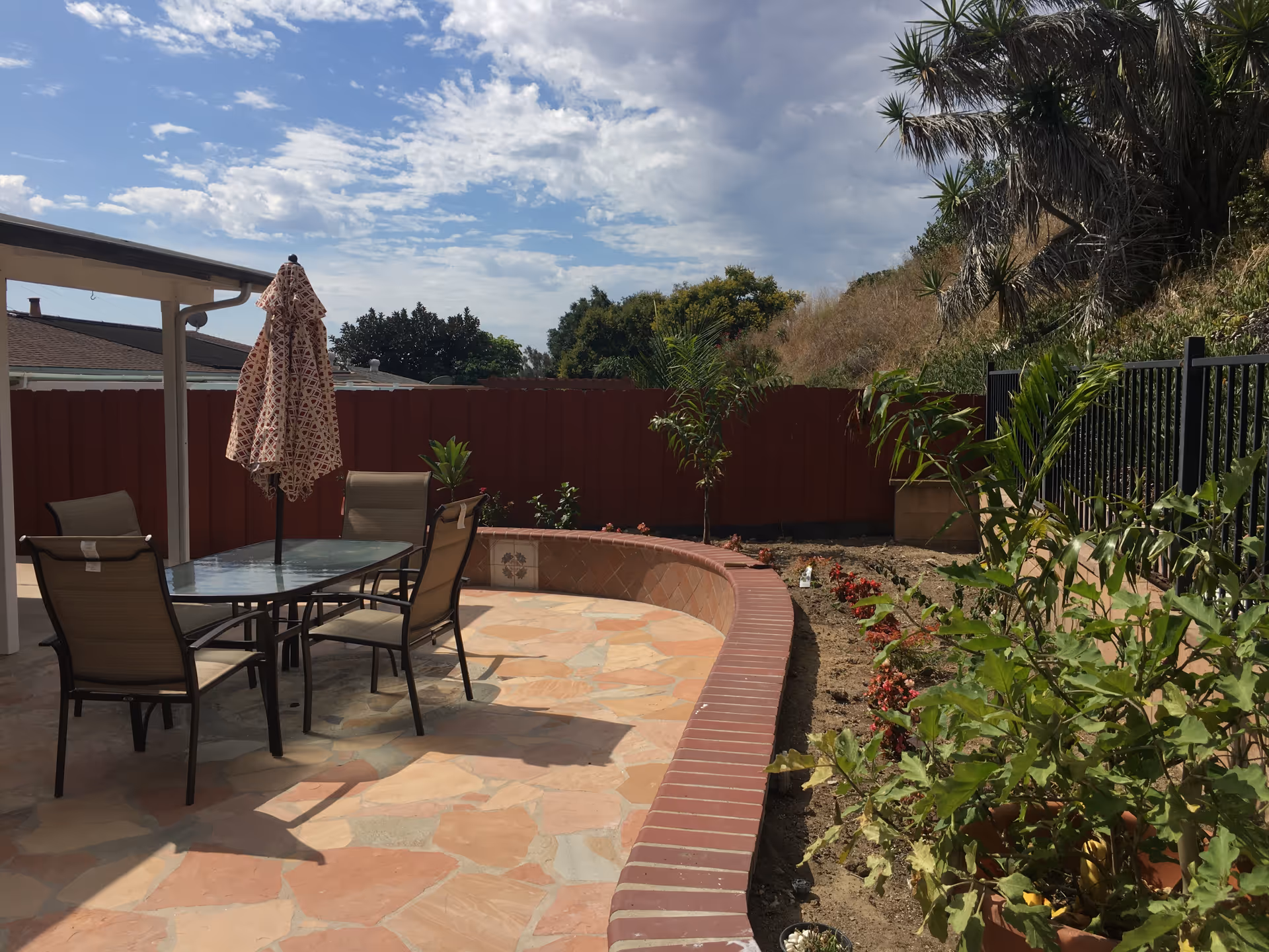 Outdoor patio with glass table, chairs, umbrella, and garden beside a red fence.