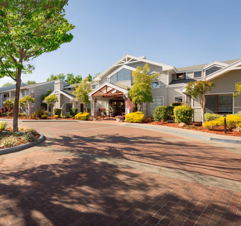 Exterior view of Cogir of Sonoma senior living facility showing a large building with multiple windows, a covered entrance with an American flag, surrounded by well-maintained landscaping including trees, bushes, and flower beds under a clear blue sky.