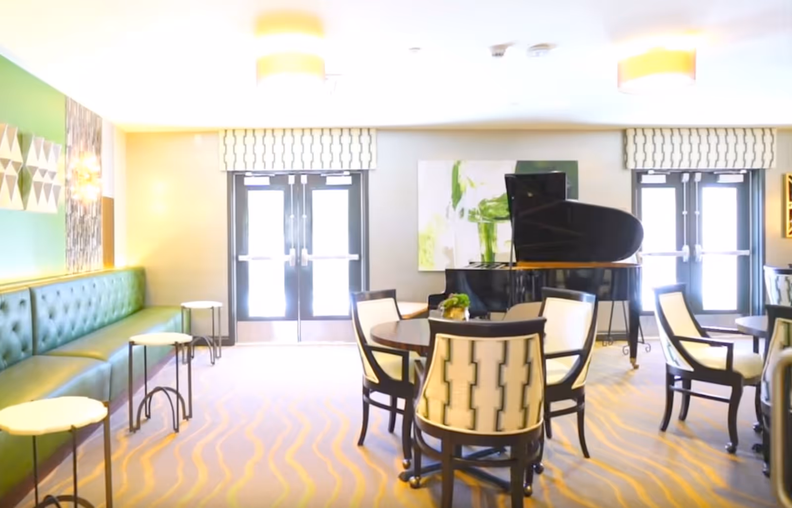 Bright communal lounge with a black grand piano, round seating tables and a green tufted bench near double doors.