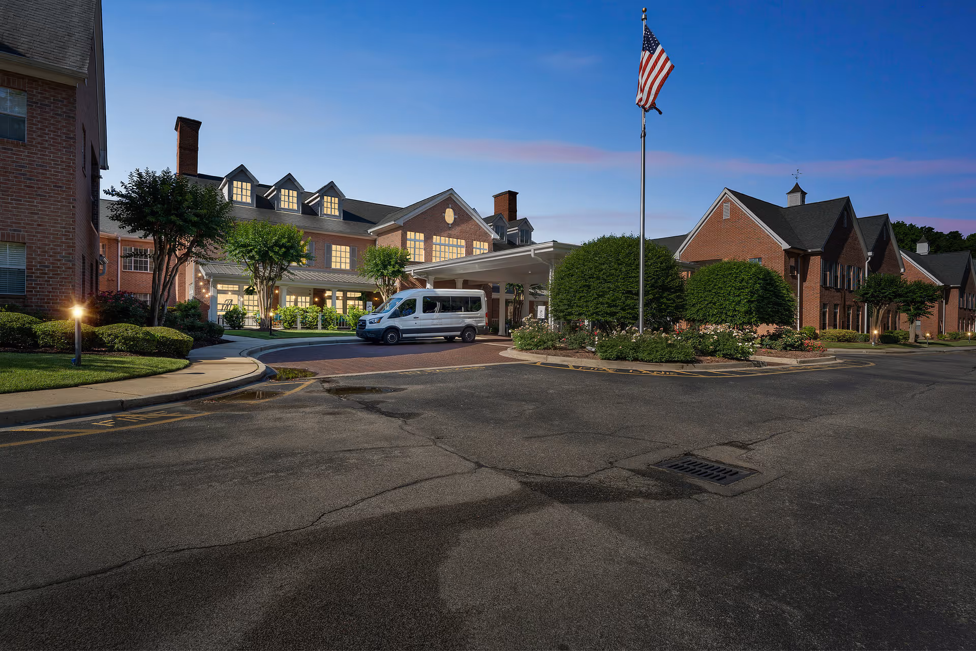 Exterior view of Town Village Audubon Park senior living facility during dusk, showing a large brick building with lit windows, a covered entrance with a white van parked underneath, landscaped bushes and trees, and an American flag on a flagpole.