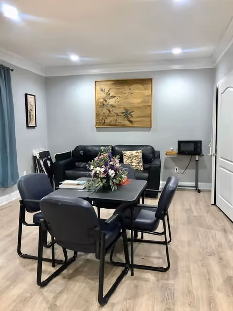 Communal living area with a table and four chairs, a floral centerpiece, a black leather sofa, wall art, and a microwave on a small side table.