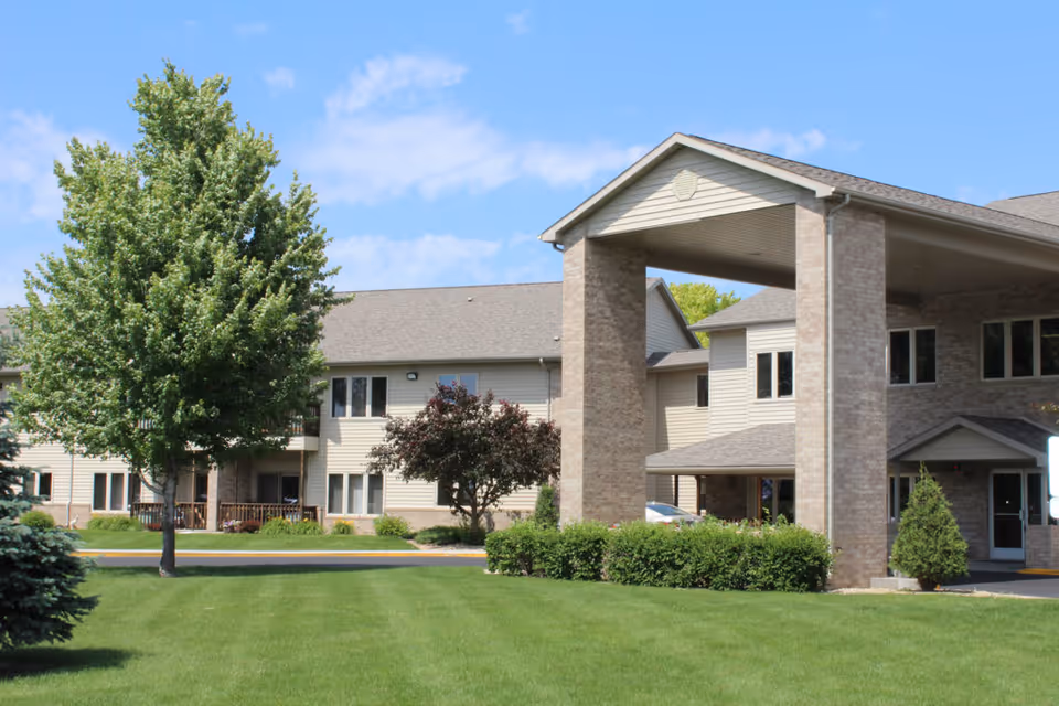 Exterior view of a senior living facility named Countryside Living with a large covered entrance, beige siding, brick pillars, multiple windows, green lawn, and trees under a blue sky with some clouds.