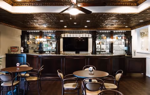Interior view of a senior living facility lounge area featuring a dark wooden bar with glass shelves displaying various items, a large flat-screen TV mounted on the wall behind the bar, round tables with chairs arranged in front, and an ornate decorative ceiling with a ceiling fan and lights.