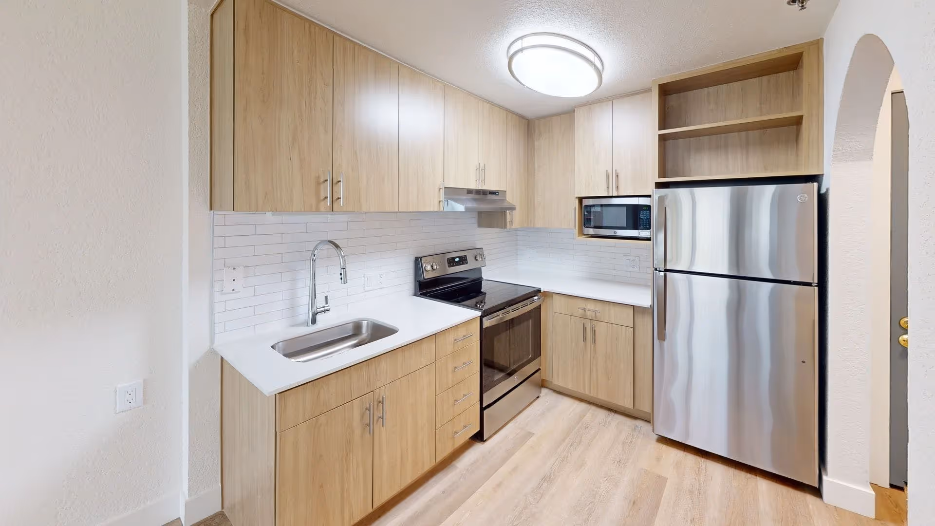 Small modern kitchen with light wood cabinets, stainless steel refrigerator and stove, a sink, microwave, and white countertops.