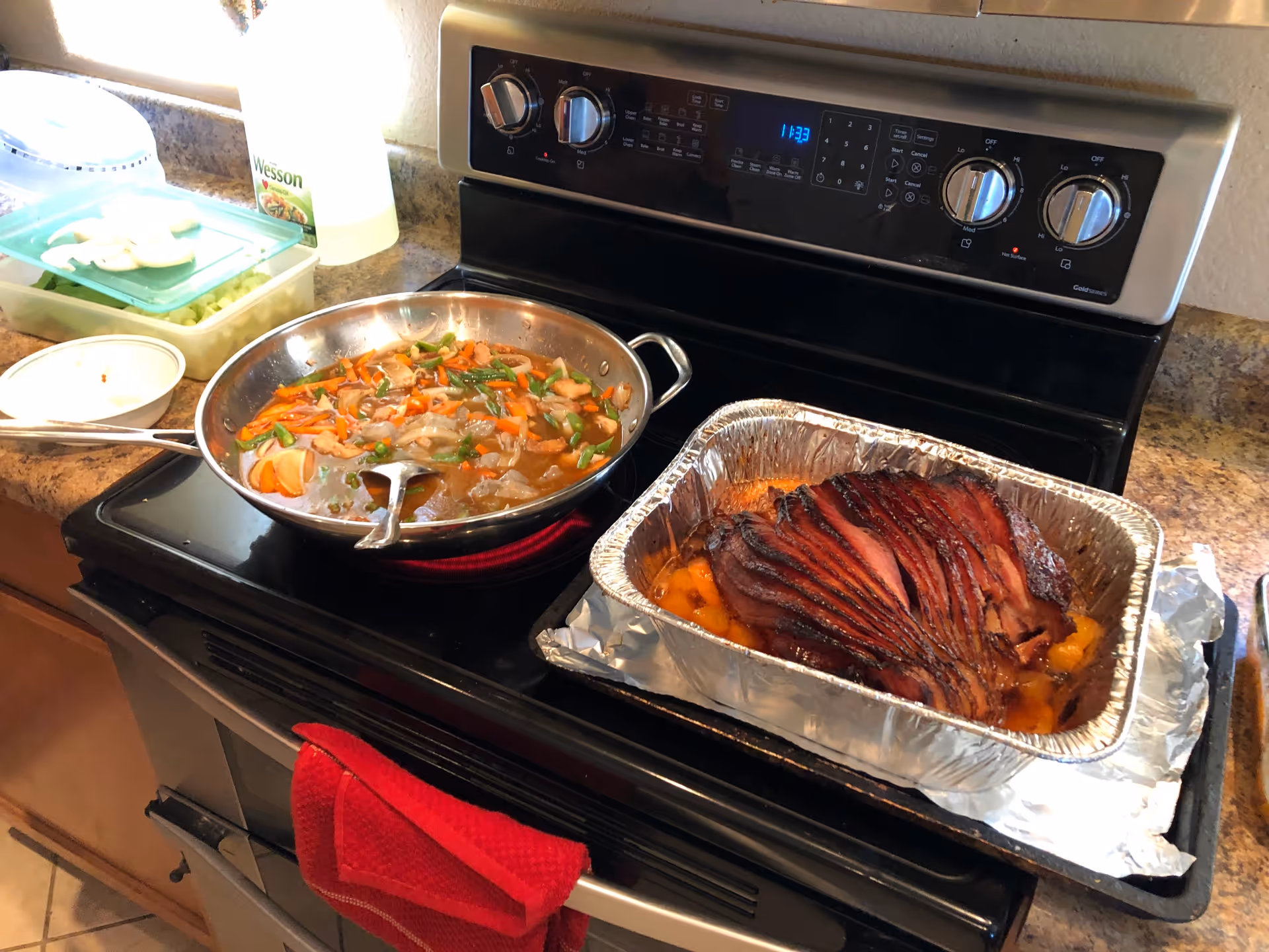 A kitchen stove with a frying pan containing cooked vegetables and a foil tray with sliced cooked ham and peaches. A red kitchen towel hangs on the oven handle, and a container with chopped vegetables and a bottle of Wesson oil are on the countertop.
