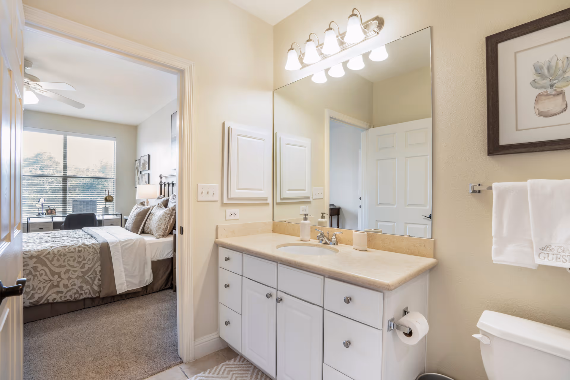 Bathroom vanity with sink and large mirror, an open door revealing a decorated bedroom.