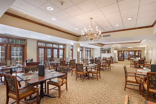 A spacious dining room with multiple tables and chairs arranged neatly. The room features large windows with wooden frames allowing natural light to enter. Elegant chandeliers hang from the ceiling, and the carpet has a subtle pattern. The tables are set with placemats, menus, and some dining utensils.