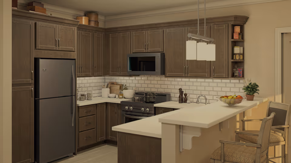 Modern kitchen with dark wood cabinets, a stainless steel refrigerator, microwave, and stove. The kitchen features white countertops and a white subway tile backsplash. There is a breakfast bar with two cushioned chairs, a bowl of fruit, and a small potted plant. Overhead lighting with three rectangular pendant lights hangs above the bar.