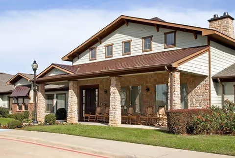 Exterior view of a single-story assisted living facility building with stone and siding walls, a covered porch with rocking chairs, a lamppost, and well-maintained landscaping including bushes and grass under a clear sky.
