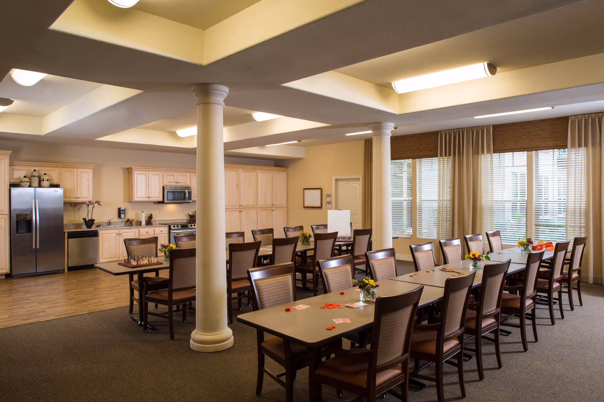 A spacious dining room with two long tables surrounded by wooden chairs. The tables have small flower arrangements and playing cards with red chips scattered on them. In the background, there is a kitchen area with light wood cabinets, a stainless steel refrigerator, dishwasher, microwave, and stove. Large windows with beige curtains allow natural light to fill the room.