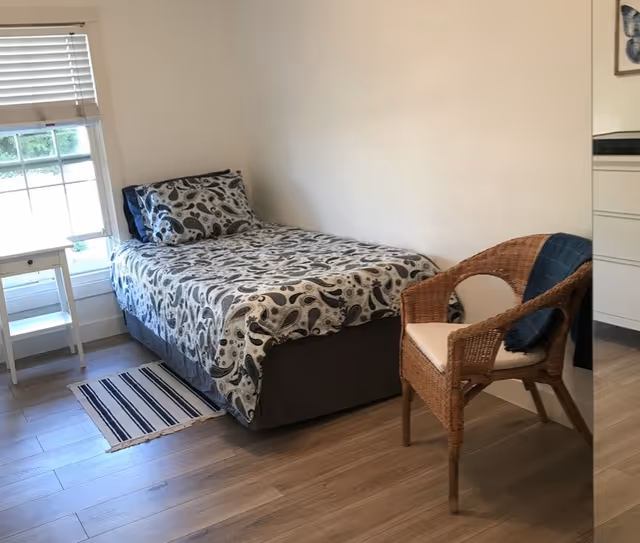 A small bedroom with a single bed covered in a patterned bedspread and matching pillow. Next to the bed is a small white side table near a window with blinds. A wicker chair with a cushion and a blue throw is placed beside the bed. The floor is wooden and there is a small striped rug near the bed.