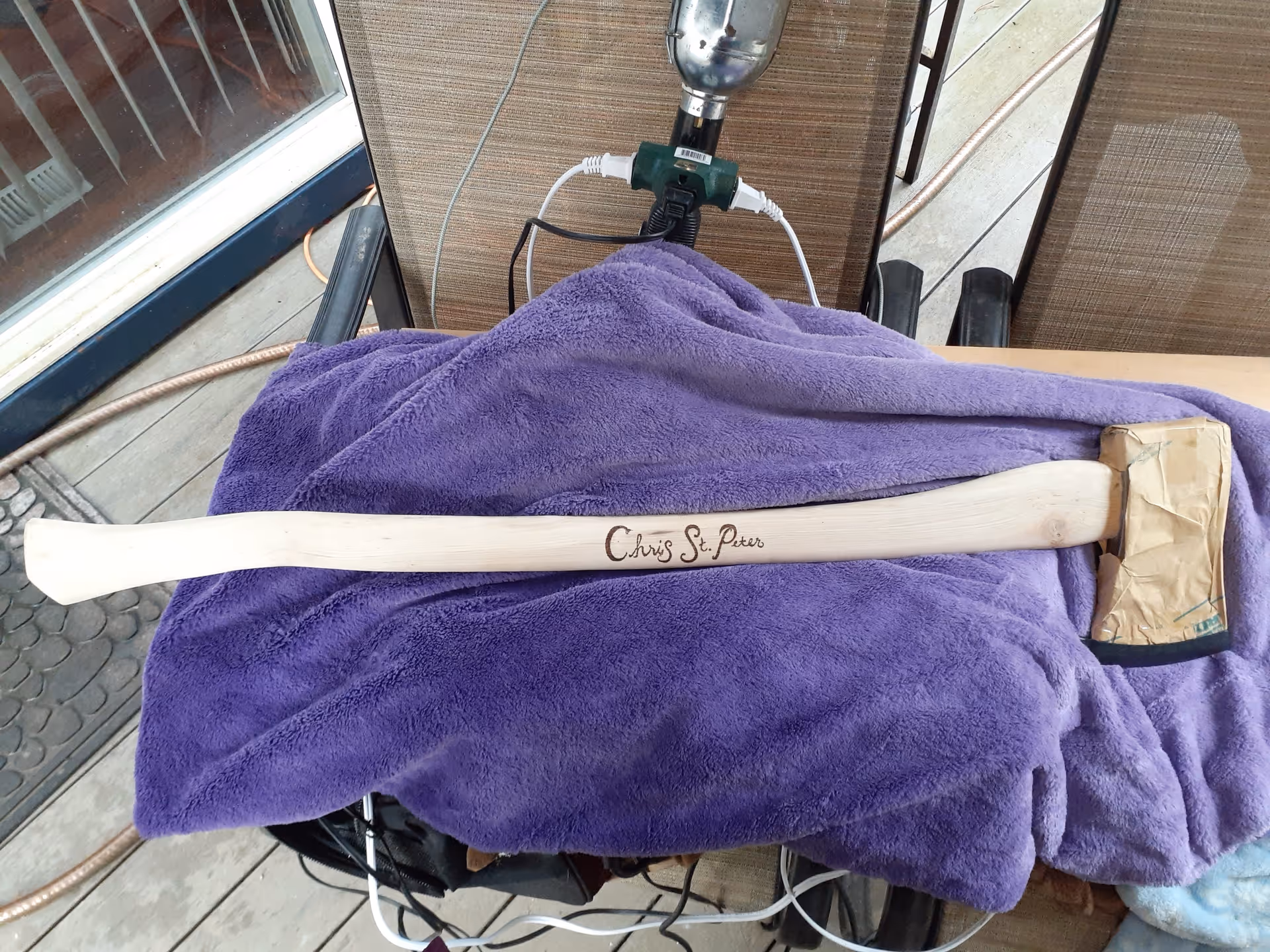 A wooden-handled axe engraved "Chris St. Peter" resting on a purple towel on a porch deck.