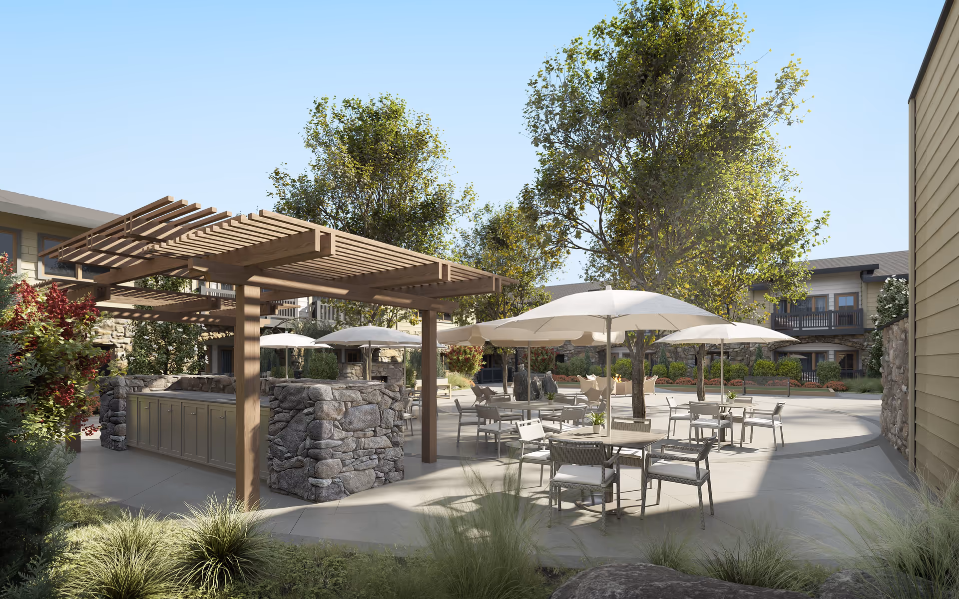 Outdoor patio area at Hearthstone Lodge Senior Residences featuring multiple tables with chairs and large white umbrellas. There is a wooden pergola structure with stone pillars and cabinetry underneath. The area is surrounded by trees, shrubs, and a two-story building with balconies in the background under a clear blue sky.