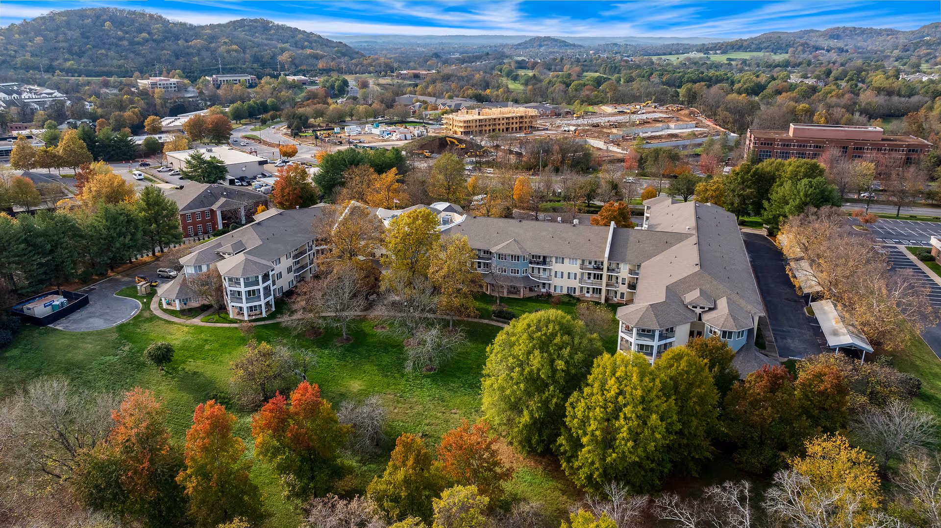 Aerial view of Manor at Steeplechase, a senior living community surrounded by green lawns and trees with autumn foliage, with hills and other buildings in the background under a partly cloudy sky.
