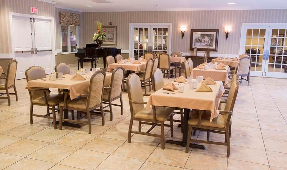A dining room with several tables covered in beige tablecloths, each set with folded napkins, cups, and utensils. The room has tiled floors, beige striped wallpaper, framed artwork on the walls, and a black grand piano in the corner. There are multiple chairs around each table and double doors with glass panes on one side.