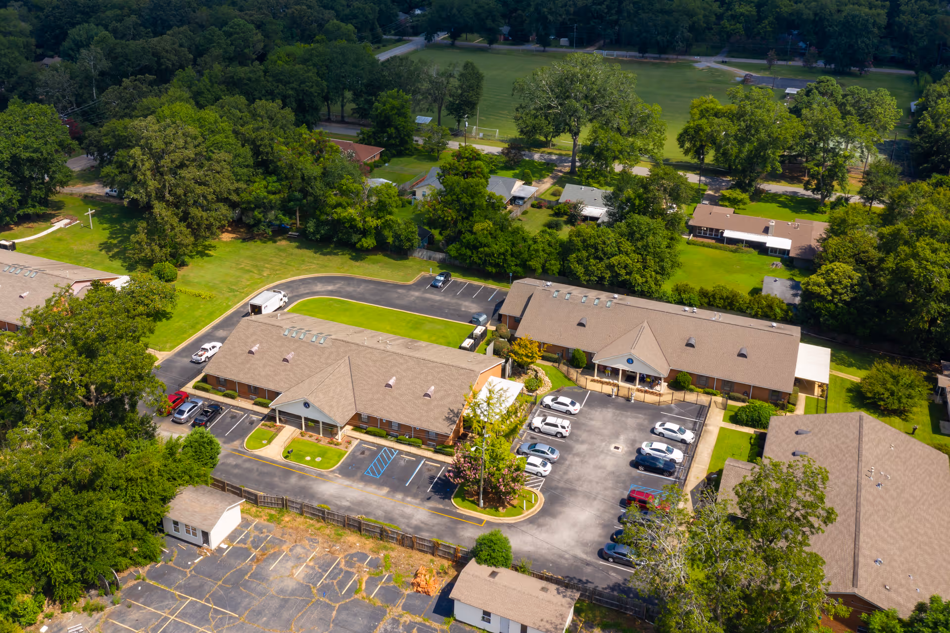 Aerial view of a senior living facility with multiple single-story buildings surrounded by trees and greenery. There are parking lots with several cars parked, paved roads, and well-maintained lawns. The facility is situated near a large open green field and residential houses.
