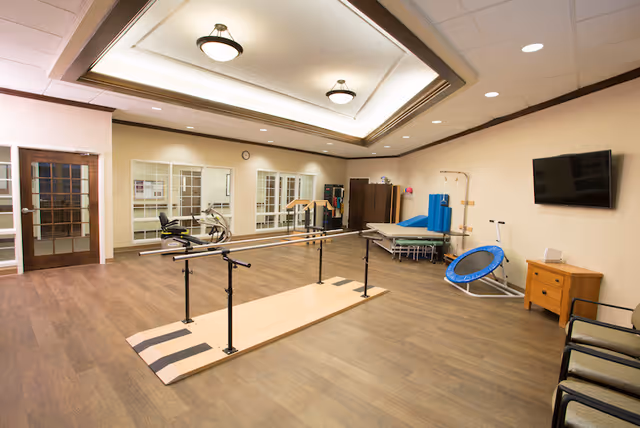 A spacious rehabilitation or physical therapy room with wooden flooring, parallel bars for walking exercises, a small trampoline, exercise equipment, a treatment table with blue cushions, chairs along the wall, a wall-mounted TV, and large mirrors on one side of the room.
