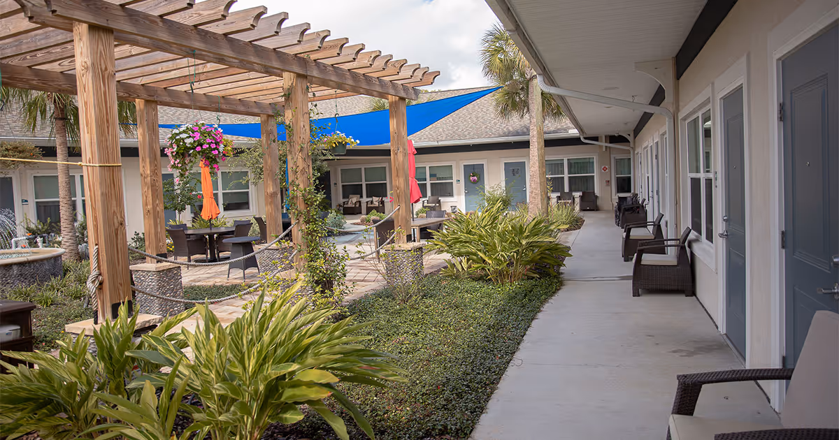 Outdoor courtyard area of an assisted living facility with a wooden pergola, hanging flower baskets, blue shade sails, patio tables with umbrellas, and surrounding greenery. There are multiple doors and windows along a covered walkway with chairs placed outside each door.