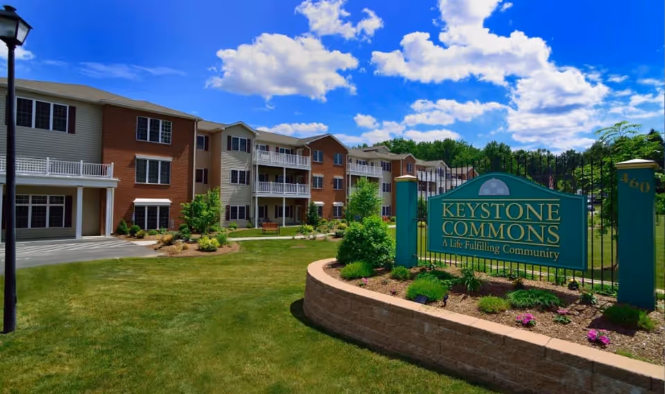 Exterior view of Keystone Commons senior living facility showing a multi-story building with balconies, well-maintained lawn, garden beds, and a large green sign with gold lettering that reads 'Keystone Commons A Life Fulfilling Community' under a bright blue sky with scattered clouds.