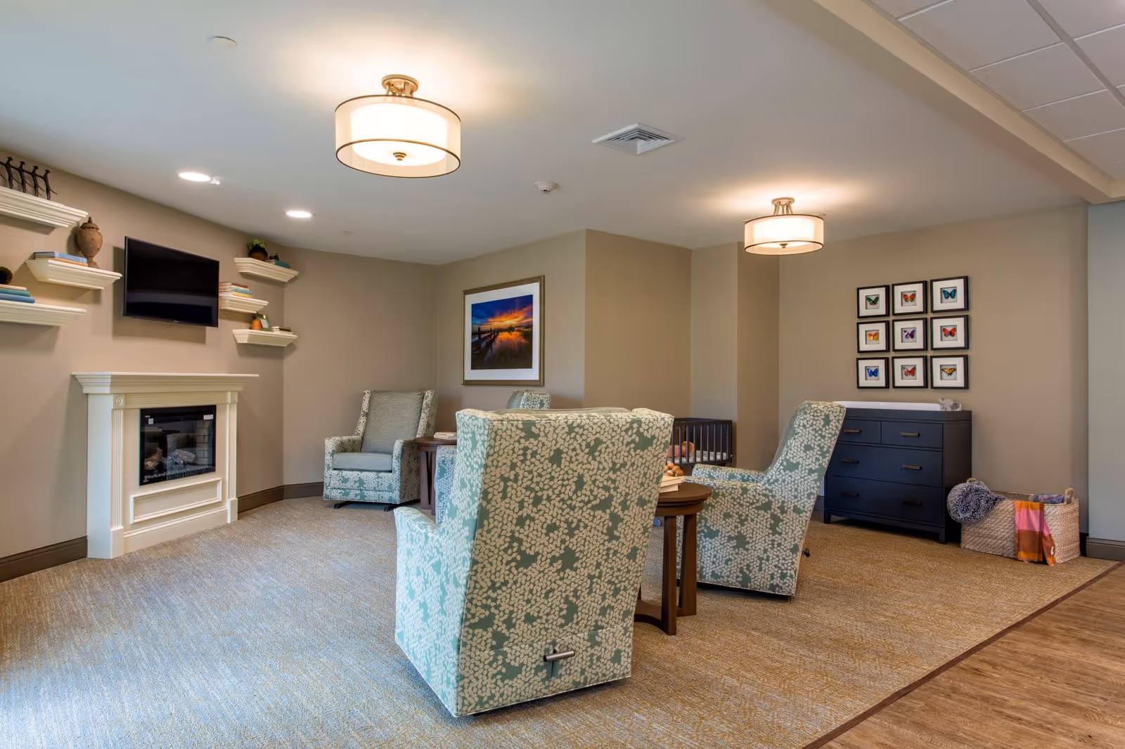 Cozy common room with patterned armchairs around a small table, a wall-mounted TV above a fireplace, and framed artwork and shelving on the walls.