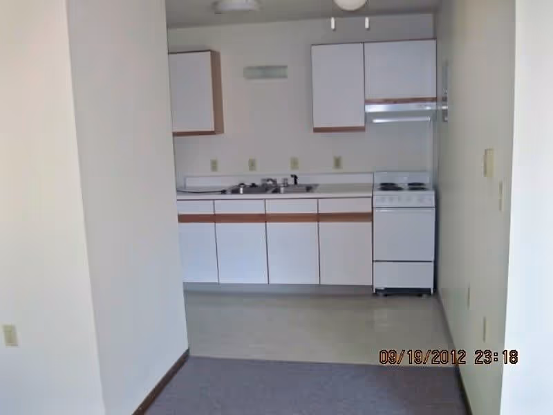 View of a small kitchen area in a senior living facility with white cabinets, a sink, and a stove. The kitchen is adjacent to a carpeted room with white walls and a beige floor in the kitchen area.