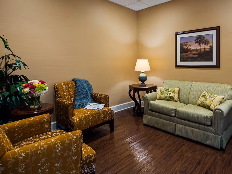 A cozy sitting area with two mustard yellow armchairs featuring a floral pattern, a green striped sofa with floral cushions, a wooden side table with a lamp, a framed landscape painting on the wall, and a small round table with a vase of colorful flowers and a green leafy plant in the corner.