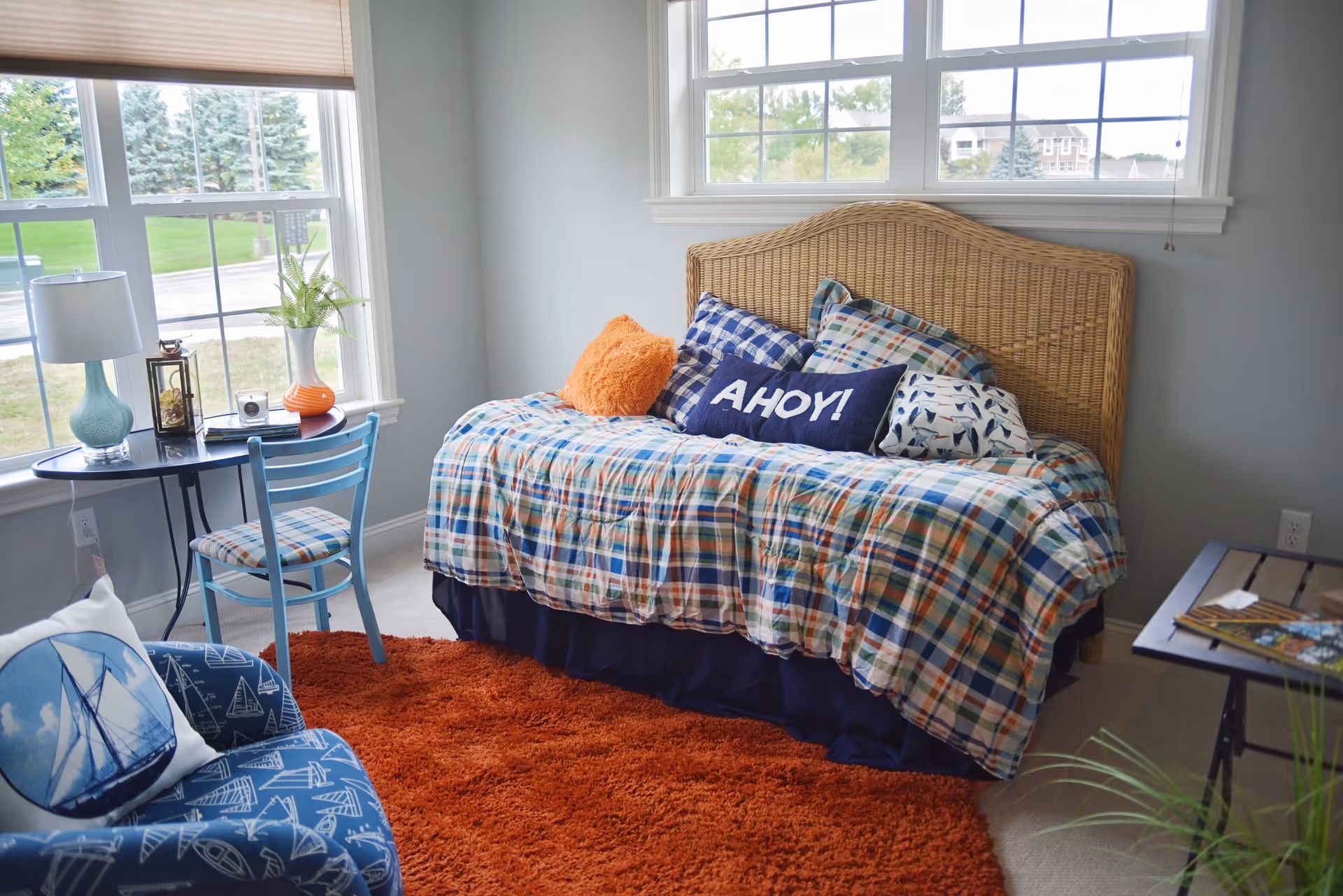 A cozy bedroom with a wicker headboard bed covered in a colorful plaid comforter and multiple pillows, including one with the word 'AHOY!'. The room has two large windows letting in natural light, a small round table with a blue chair, a lamp, and decorative items. There is also a blue armchair with a sailboat pillow and an orange shag rug on the floor.