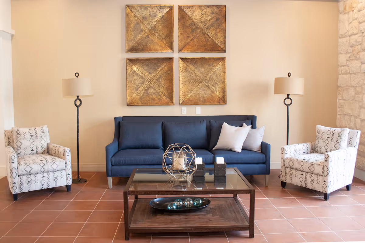 A furnished living room with a blue sofa, two patterned armchairs, floor lamps, a glass-top coffee table, and four gold decorative wall panels.