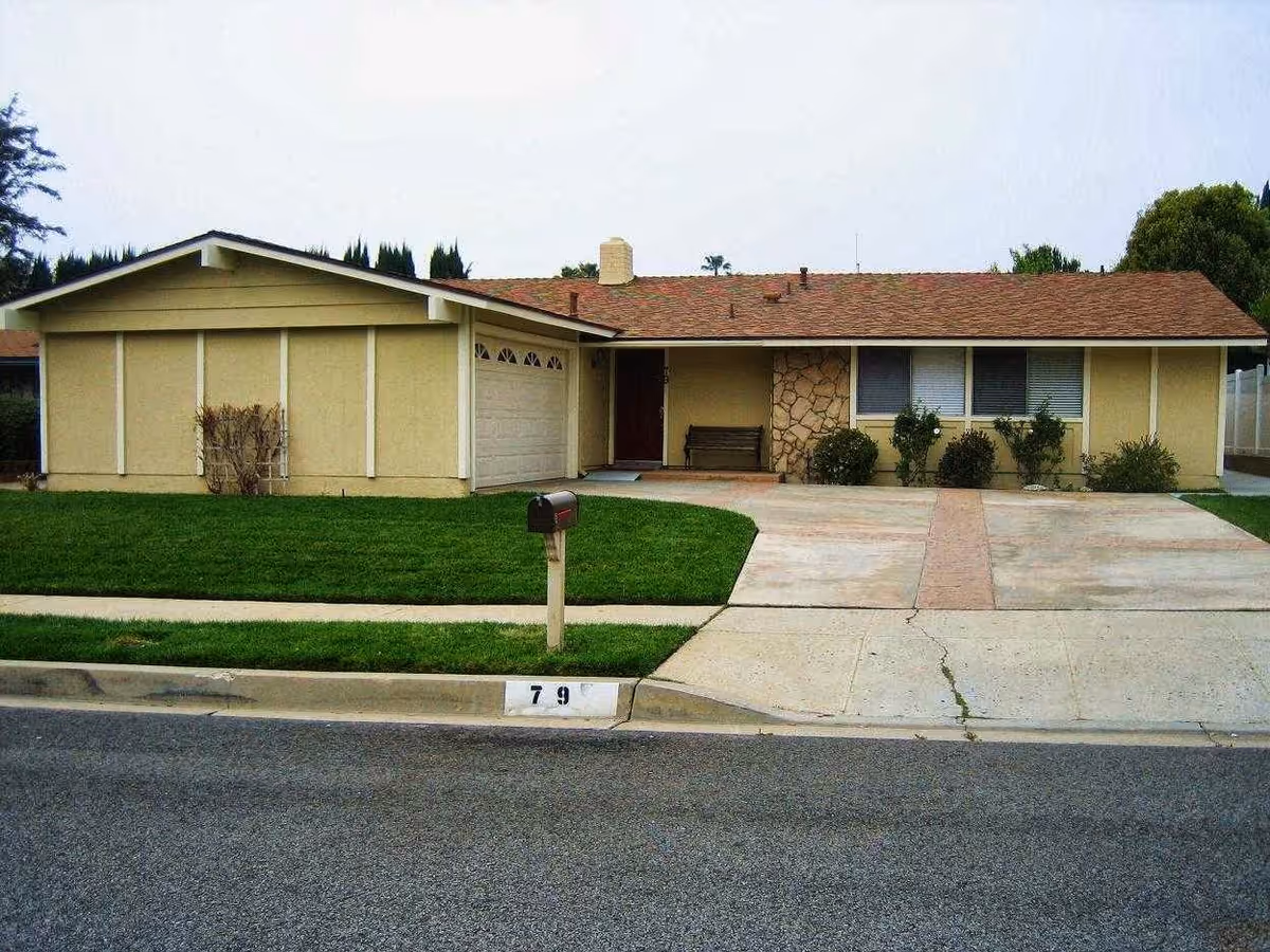 Single-story residential house with a beige exterior, a two-car garage, a driveway, a small front lawn with a mailbox, and some bushes near the front windows under an overcast sky.