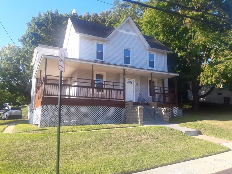 A two-story white residential building with a brown wooden porch railing and steps leading up to the front door. The house is surrounded by green grass and trees, with a 'No Stopping' sign visible in the front yard.