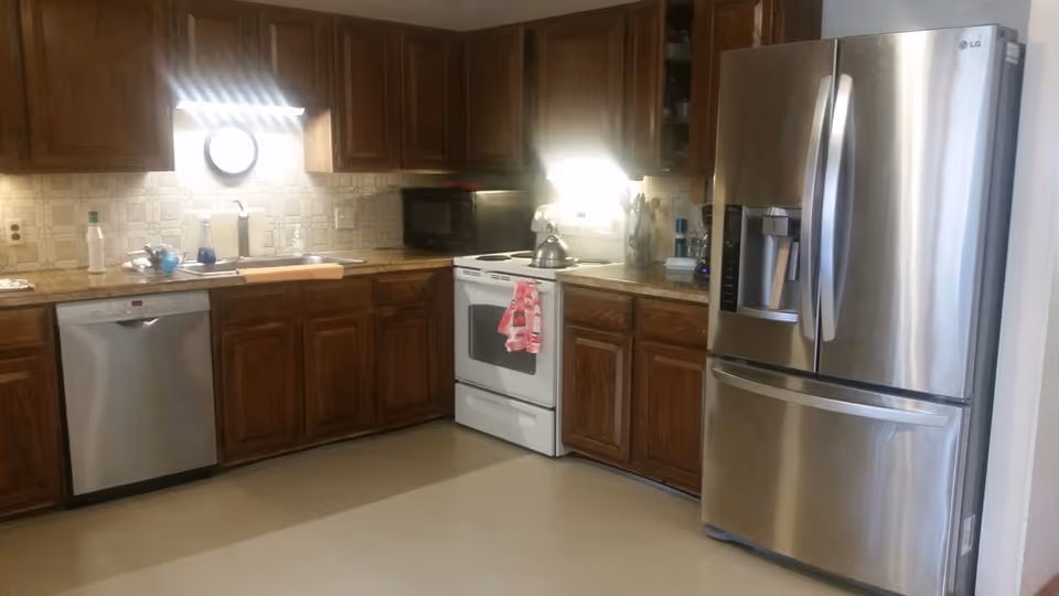 A kitchen with dark wooden cabinets, a stainless steel French-door refrigerator and dishwasher, a white stove, and countertops with a microwave and sink.