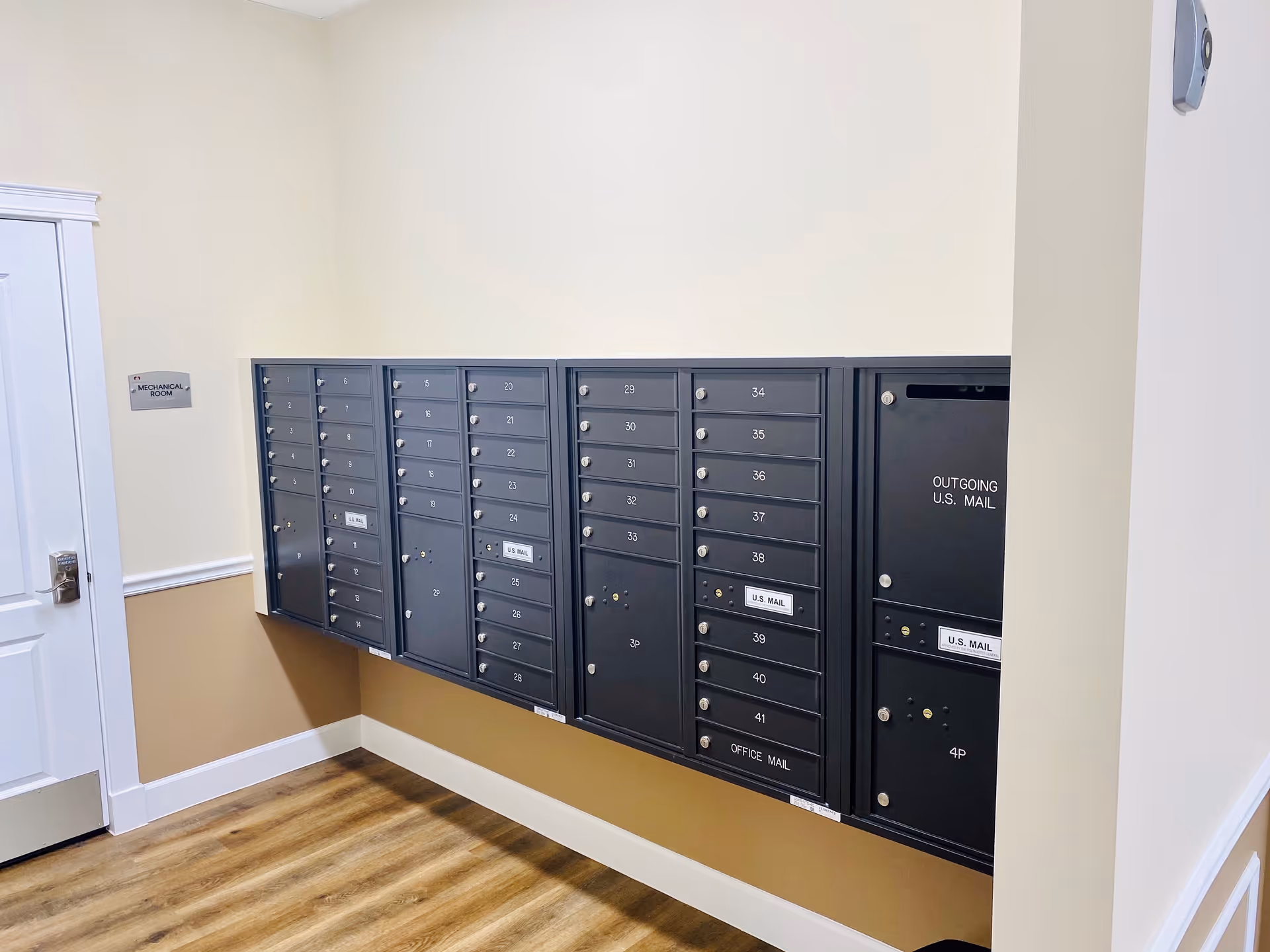 Interior view of a mailroom area with multiple black mailboxes mounted on a beige and white wall. There is a white door labeled 'Mechanical Room' on the left side and a wooden floor beneath.