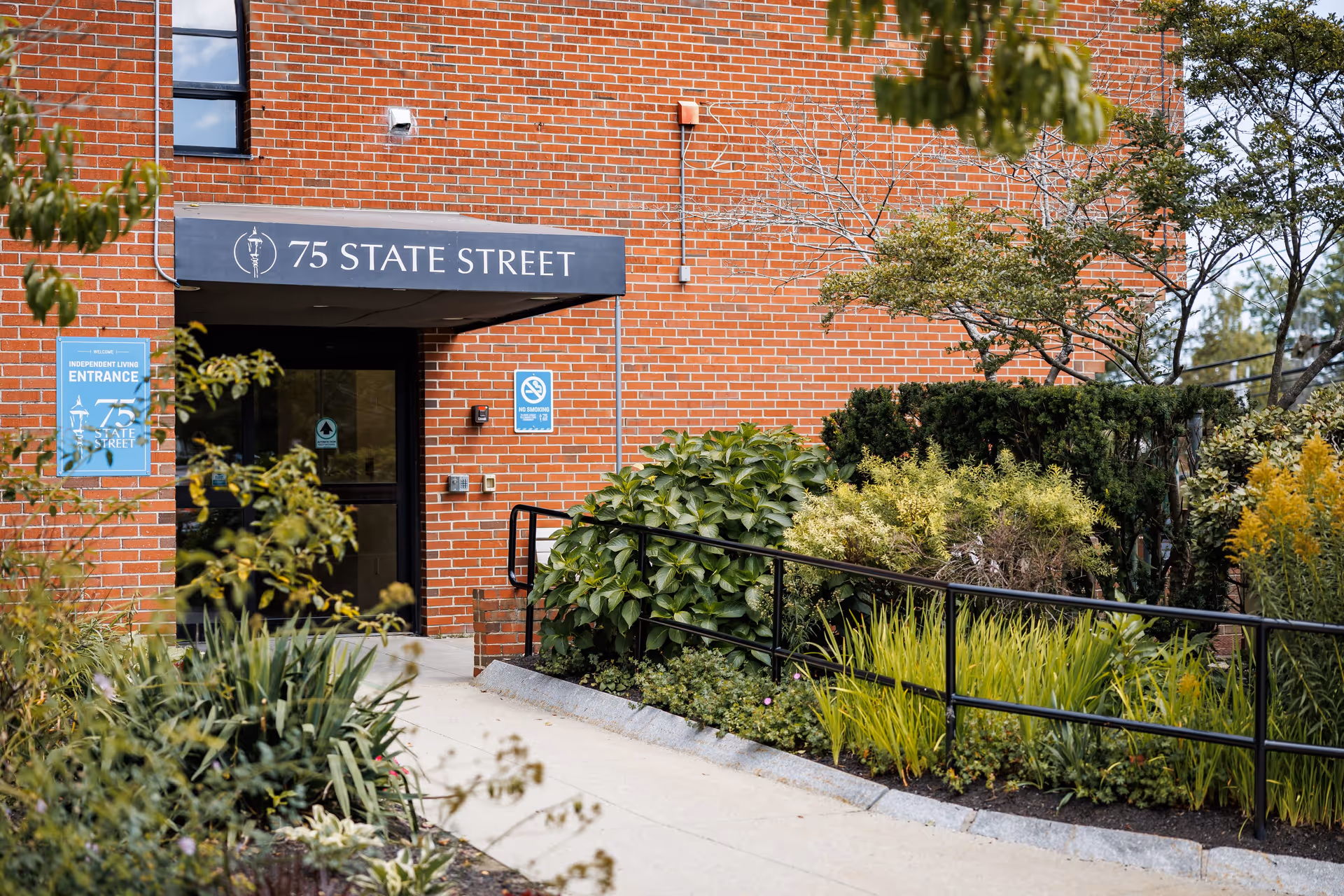 Entrance to a brick building with a black awning displaying the text '75 State Street'. There is a sidewalk leading to the door, surrounded by green shrubs and plants. A blue sign on the left side of the entrance indicates 'Independent Living Entrance'.