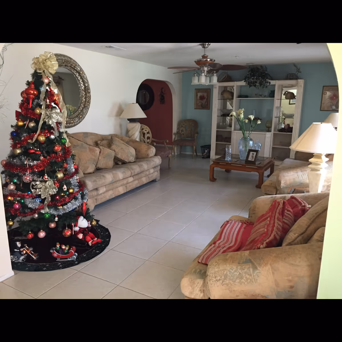 A cozy living room decorated for Christmas with a decorated Christmas tree on the left side. The room features beige sofas with patterned cushions, a wooden coffee table with a vase of flowers and framed photo, a white display cabinet with decorative items, a ceiling fan with lights, and a large round mirror on the wall.