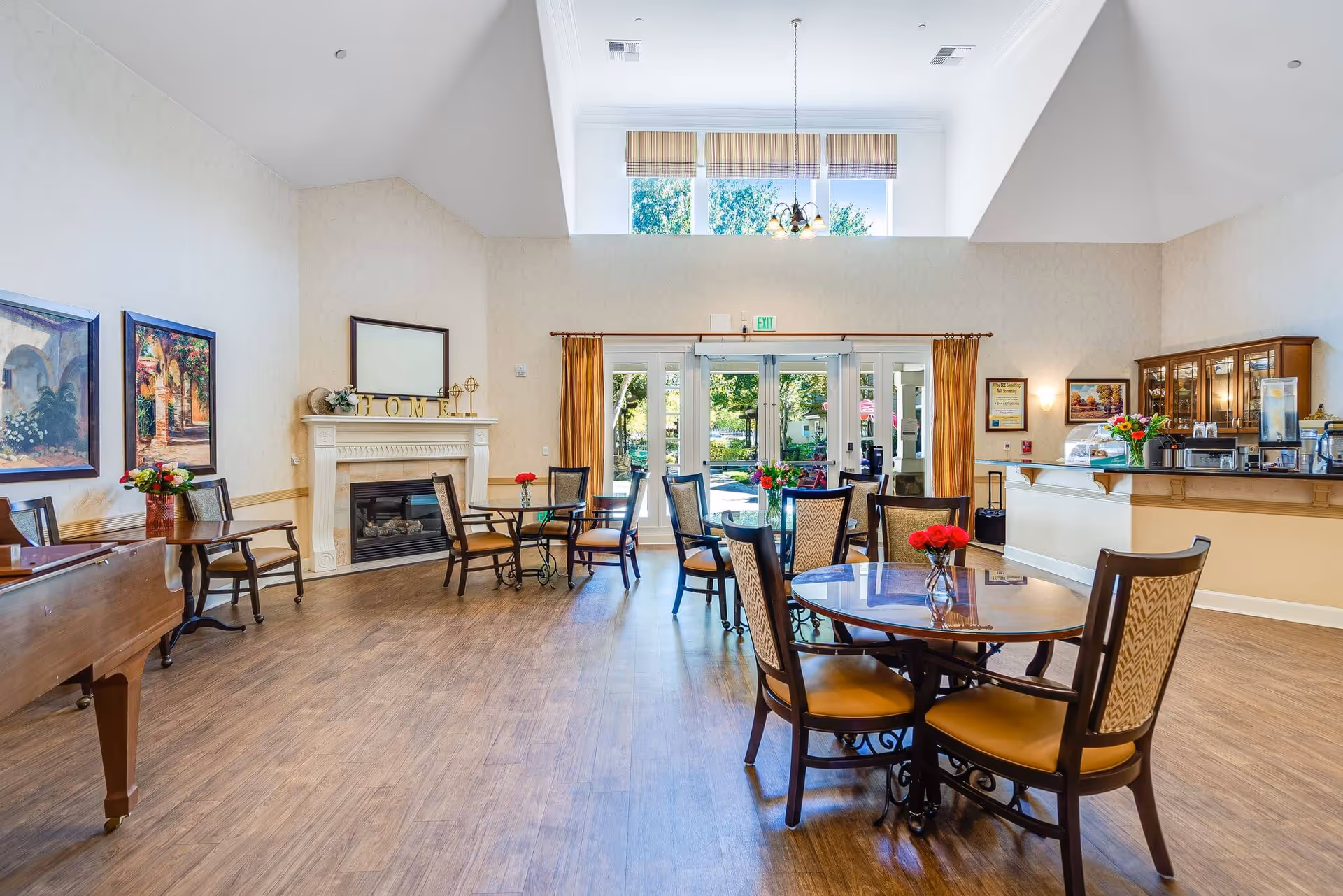 Bright communal dining area with round tables and chairs, a fireplace, high windows, and a service counter.
