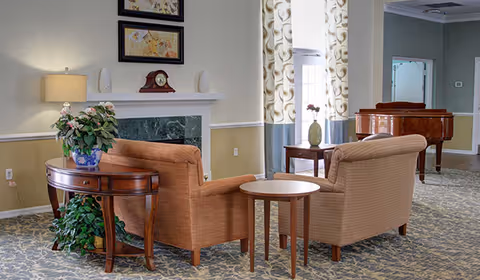 A cozy senior living common area with two upholstered armchairs facing a small round table. Behind the chairs is a wooden side table with a potted plant. The room features a fireplace with a clock and decorative items on the mantel, framed artwork above, patterned carpet, and large windows with patterned curtains. A grand piano is visible in the background.