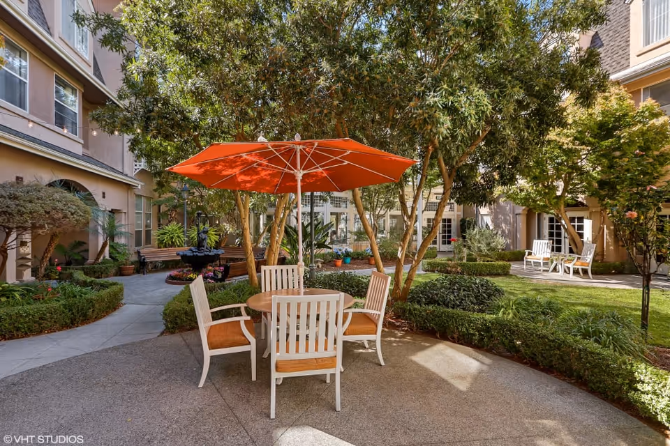 Outdoor courtyard area at Sunshine Villa, A Merrill Gardens Community, featuring a round table with four white chairs and an orange umbrella. The courtyard is surrounded by trees, bushes, and well-maintained landscaping with a pathway and benches visible in the background.