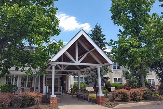 Entrance of a senior living facility with a covered driveway supported by white beams and brick pillars, surrounded by trees, shrubs, and flowers under a partly cloudy sky.