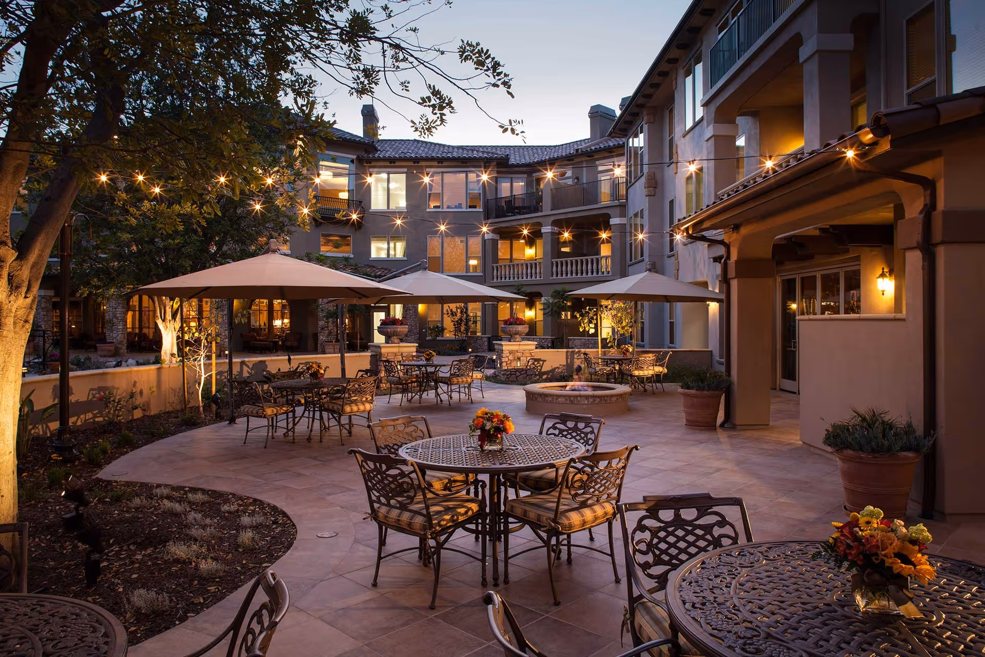 A landscaped courtyard patio at dusk with metal tables and chairs, umbrellas, string lights, and a surrounding multi-story building.