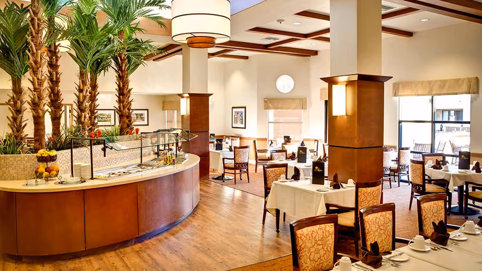 A spacious dining room with multiple tables covered in white tablecloths, each set with cups, plates, and folded napkins. The room features wooden beams on the ceiling, large windows allowing natural light, and a curved buffet counter with a fruit display and plates. Tall indoor palm trees add greenery to the space.
