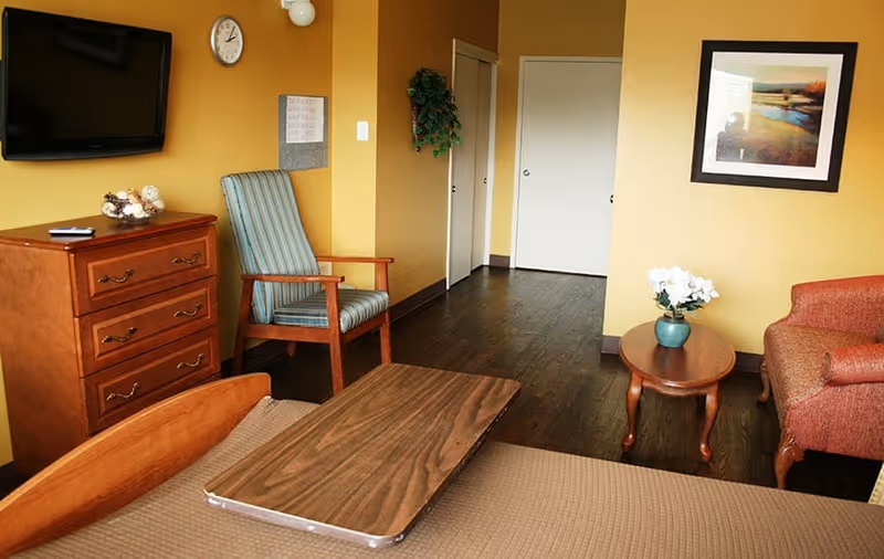A cozy rehab bedroom featuring a bed with an overbed table, wooden dresser and wall-mounted TV, armchairs, a side table with flowers, and warm yellow walls.