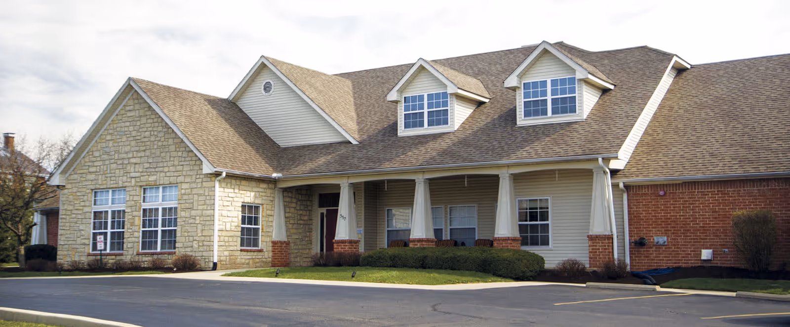 Exterior view of a single-story building with a combination of stone, brick, and siding on the facade. The building has a sloped roof with two dormer windows and a covered entrance supported by columns. There is a paved driveway and some landscaping with bushes and grass in front.
