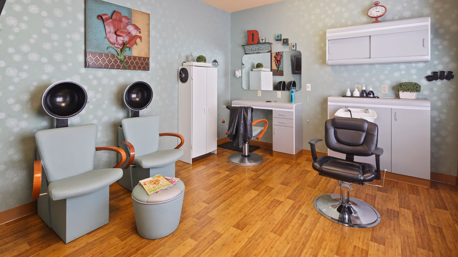 Interior of a hair salon room with two vintage hair drying chairs on the left, a small ottoman with magazines, a white cabinet, a styling station with a mirror and chair, and a black salon chair in front of a counter with a sink and cabinets. The walls are light green with a subtle floral pattern, and there is a painting of a pink flower on the wall.
