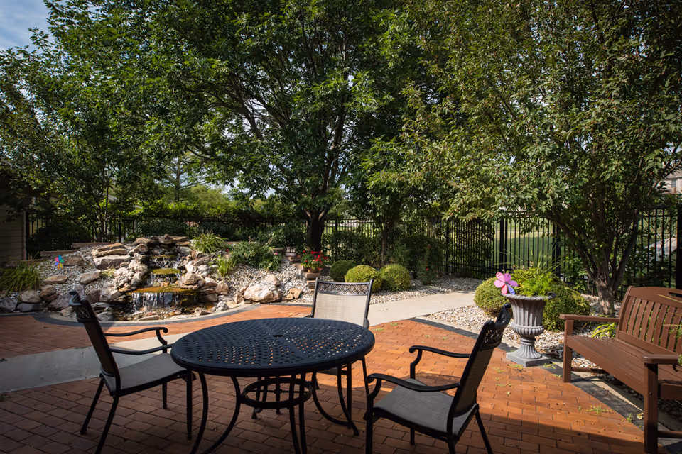 Outdoor patio area with a round metal table and three chairs on a brick surface, surrounded by trees, bushes, and a small rock waterfall feature in the background.