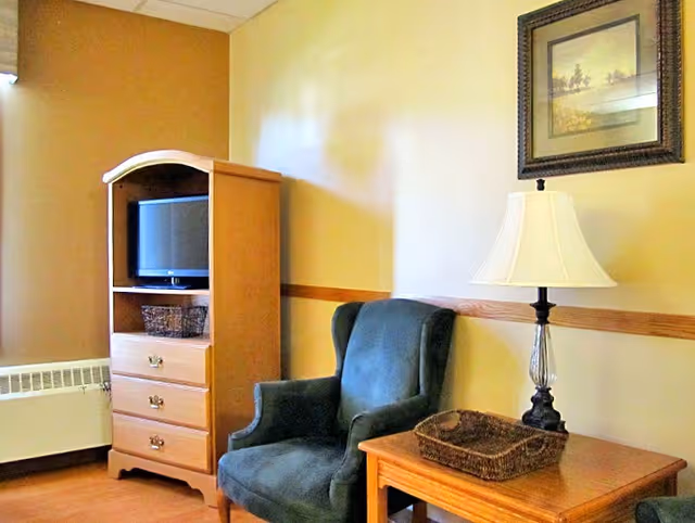 A cozy corner of a living room in a senior living facility featuring a dark green upholstered armchair, a wooden side table with a wicker basket and a lamp with a white shade, a wooden cabinet with a small television and a wicker basket on the shelf, and a framed picture hanging on the beige wall.
